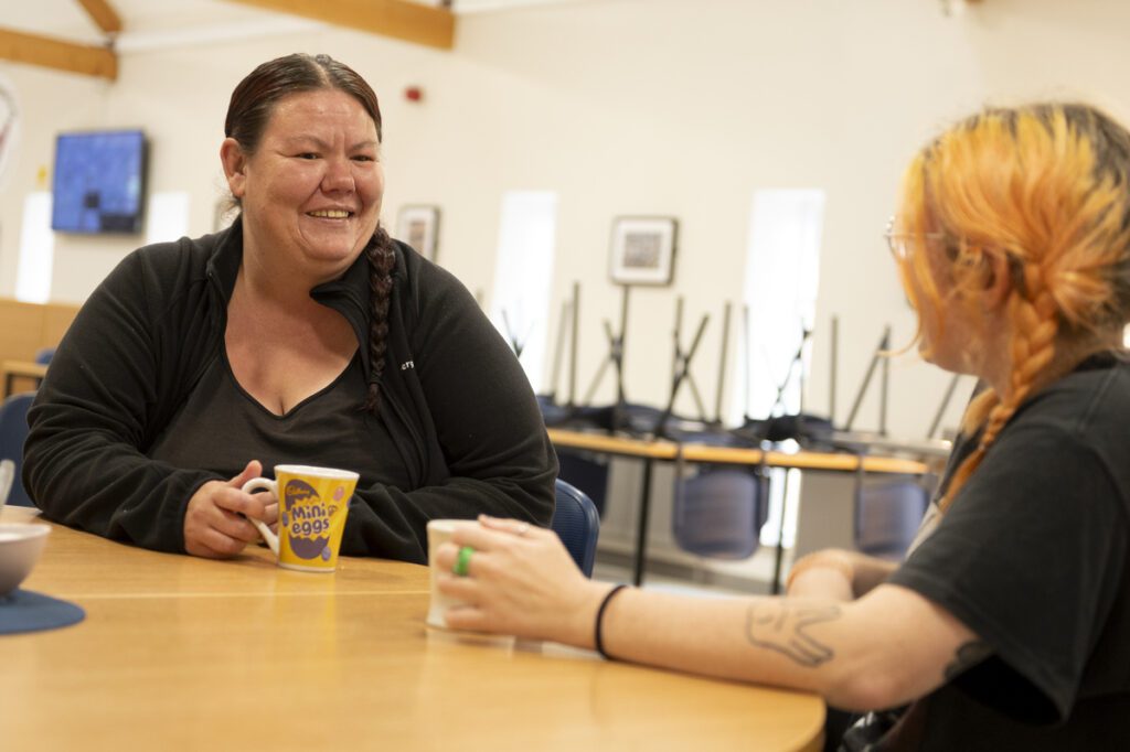 Woman with mug speaking to another woman at a table